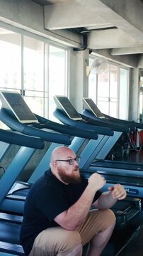 Person sitting on a treadmill with head in hands in a gym, beside a row of empty machines, sunlight streaming through large windows in an industrial-style workout space.