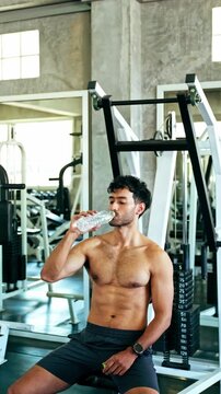 Shirtless athlete rests on a gym machine, sipping water during a workout in a modern fitness center