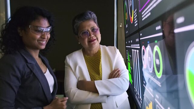 Two professionals examine a large digital dashboard with charts and metrics in a dim office, discussing performance insights.