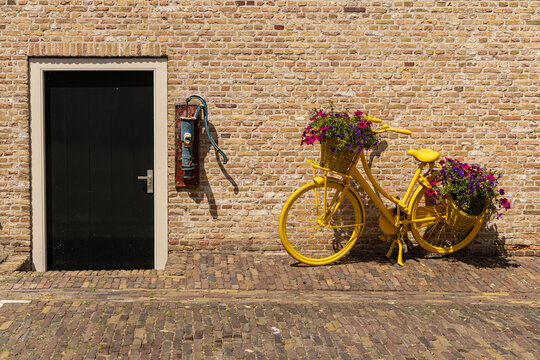 View of a yellow bicycle with flower baskets parked against a brick wall next to a black door and an old water pump in Heusden, North Brabant, Netherlands.