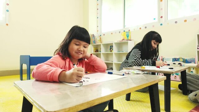 Two children draw with markers at small desks in a bright classroom, focused on creative schoolwork amid shelves and windows.