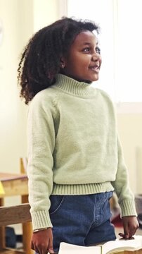 A young student stands by a desk in a bright classroom, speaking confidently beside an open book, wearing a light turtleneck sweater and jeans.