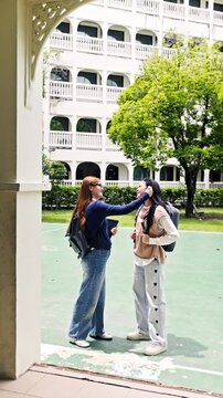 Two students with backpacks stand in a school courtyard; one uses a handheld device to check the other's temperature near a white arched building and trees.