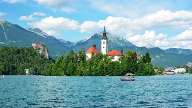Scenic view of the iconic Church of the Assumption on Bled Island in Lake Bled, Slovenia, with unrecognizable people boating beneath a blue sky with scattered clouds.