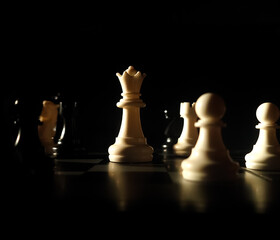 Low-key close-up photograph of a white chess queen illuminated by dramatic directional lighting on a chessboard, surrounded by blurred chess pieces against a dark black background. © Reza