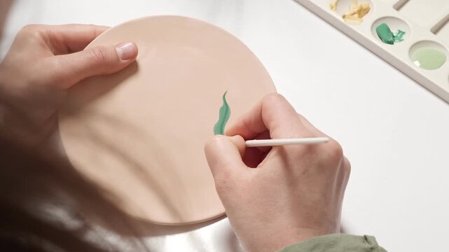 4k video Process of Drawing and Painting on a Ceramic Plate in Slow Motion. Artist painting a ceramic plate. An artisan uses a fine brush to apply green underglaze, creating botanical artwork