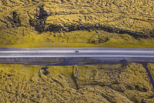View of a lone car driving along a straight asphalt road cutting through a vast, moss-covered volcanic landscape in Iceland.