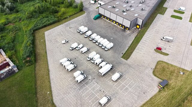 Aerial view of parked delivery trucks in a logistics company yard, showcasing organized rows of vehicles and a large warehouse building in the background. Transportation concept