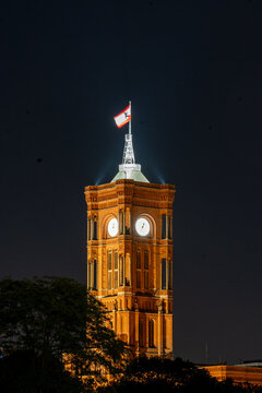 View of the illuminated clock tower of the Rotes Rathaus at night with a flag flying on top against a dark sky in Berlin, Berlin, Germany.
