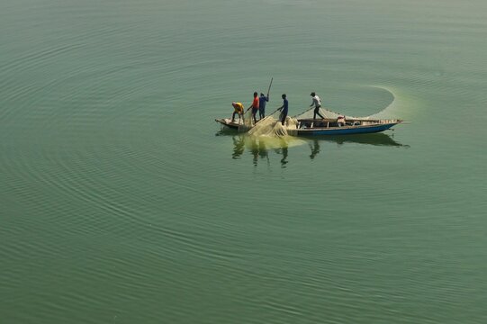 Traditional fishermen casting a net from a wooden boat on calm waters in Bangladesh.