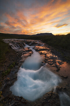 View of Bruarfoss waterfall with its characteristic bright blue glacial water flowing over dark volcanic rocks under a vibrant orange and pink sunset sky in Blaskogabyggd, Iceland.