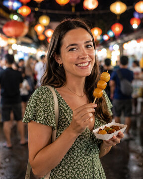 Smiling traveler tastes local street food at a lively night market, surrounded by colorful lantern bokeh and an energetic international atmosphere.