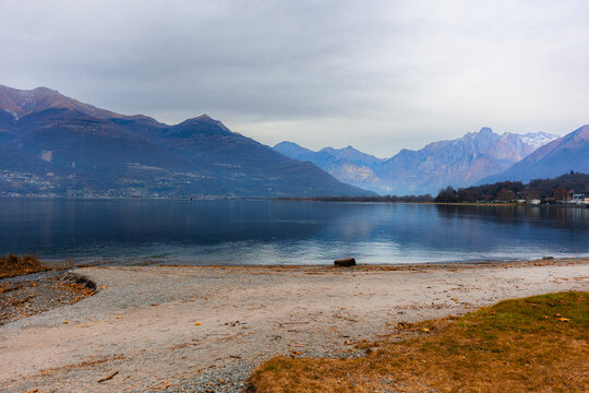 View of Lake Como in winter with calm water, a pebbled shoreline, and snow-capped mountains under an overcast sky in Colico, Lombardy, Italy.