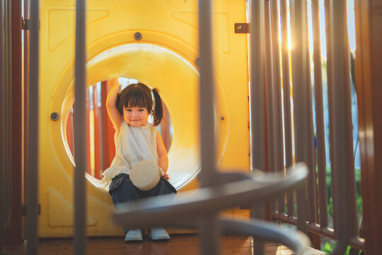 Cute Asian toddler girl sitting inside yellow tunnel playground waving hand with cheerful smile at sunset, happy little kid enjoying outdoor play activity for childhood development, family leisure