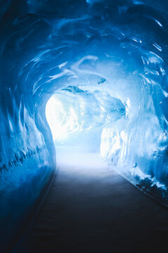 View of the illuminated ice tunnel inside the Mer de Glace glacier with smooth blue ice walls and a bright exit in Chamonix, Auvergne-Rhone-Alpes, France.