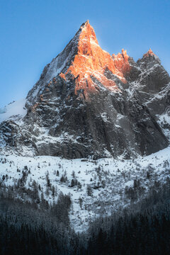 View of the snow-covered peak of Aiguille du Dru illuminated by golden sunset light above a dense pine forest in Chamonix, Auvergne-Rhone-Alpes, France.