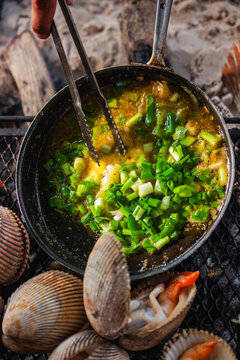 View of fresh cockles being grilled over an open fire with chopped green onions in a pan on the beach in Phu Quoc, Kien Giang, Vietnam.