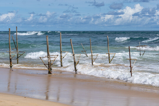 View of traditional stilt fishing poles standing in the surf at Ahangama Beach with waves crashing against the sandy shore in Galle, Southern Province, Sri Lanka.
