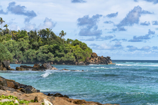 View of the turquoise waters and rocky coastline of Ahangama Beach, framed by lush tropical vegetation and palm trees under a bright blue sky in Galle, Southern Province, Sri Lanka.