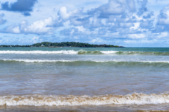 View of Weligama Beach with gentle waves rolling onto the sandy shore under a bright blue sky with scattered clouds in Weligama, Southern Province, Sri Lanka.