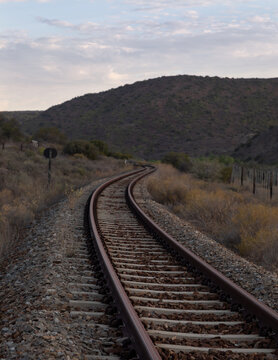 Scenic sunset view of a winding railroad track in the Karoo desert, South Africa, featuring a curved perspective of steel rails leading into a wild west badlands landscape under a pink twilight sky.