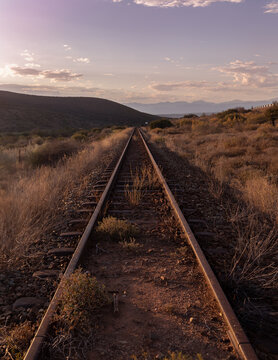 Scenic sunset view of a straight railroad track in the Karoo desert, South Africa, featuring a wide perspective of steel rails leading into a wild west badlands landscape under a pink twilight sky.