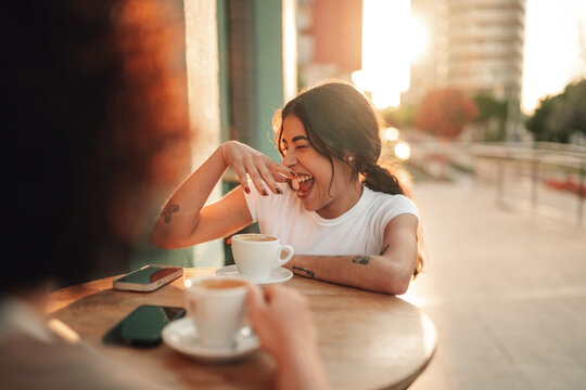 Happy young woman laughing, enjoying coffee with friend at street cafe in warm sunset light