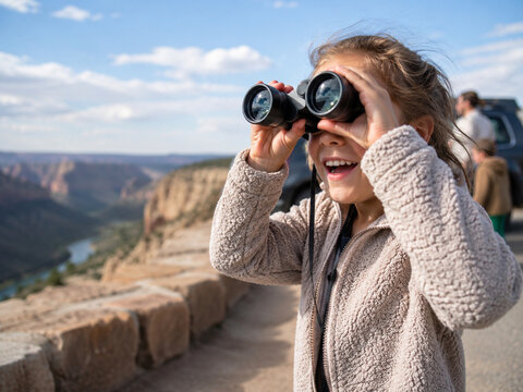 Excited child using binoculars at a canyon overlook during a family road trip, with dramatic cliffs and a parked vehicle softly blurred behind.