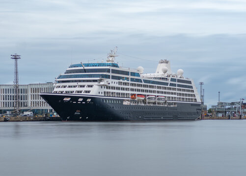 A large black and white luxury cruise ship moored at an industrial port harbor with calm water reflections under an overcast sky, port buildings visible in background