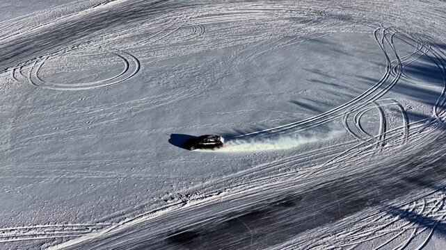 Aerial view of a black car performing a drift maneuver on a vast snow-covered field, leaving deep tire tracks in the fresh powder in Latvia.