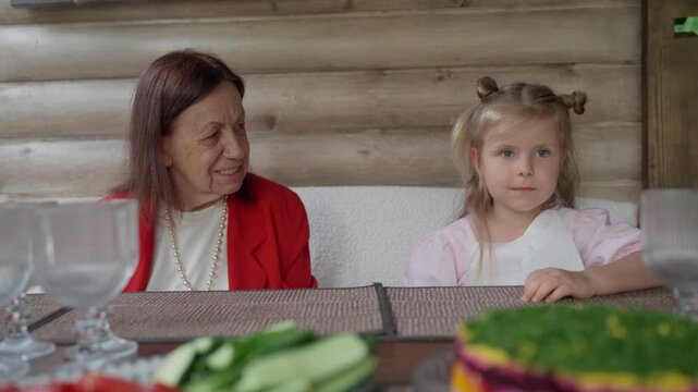 Older woman and granddaughter smiling at table. Happy little girl (4+) having fun with her senior grandmother (80+) sitting at the table while dining.
