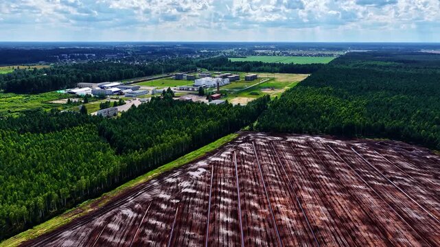 Aerial view of a harvested peat bog field adjacent to a dense forest and industrial facilities under a cloudy blue sky in Riga, Latvia.