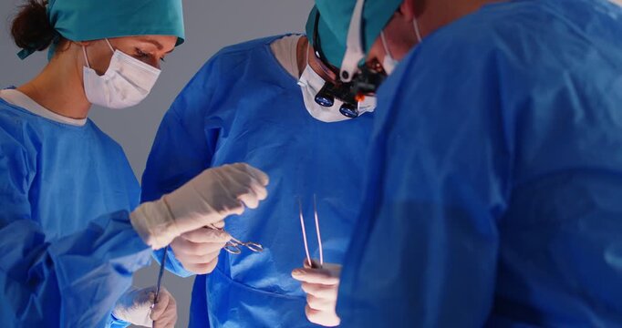 Surgeon group working, professional medical doctors surgical team performing surgery, doing operation, procedure on patient, wearing surgical gown, cap, face mask, eye loupe binocular, treat injuries