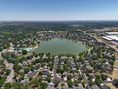 Aerial view of a residential neighborhood surrounding a central lake with suburban houses, lush trees, and clear blue sky in Thornton, Colorado, United States.