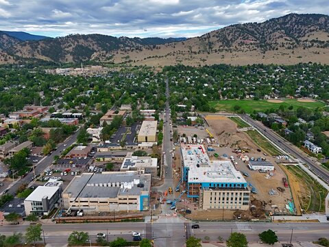 Aerial view of a construction site with modern buildings, surrounding residential neighborhoods, lush green trees, and the Rocky Mountains in the background in Boulder, Colorado, United States.