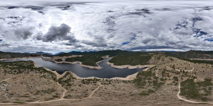 Aerial view of Gross Reservoir surrounded by hills, pine trees, and dirt trails under a cloudy sky in Boulder County, Colorado, United States.