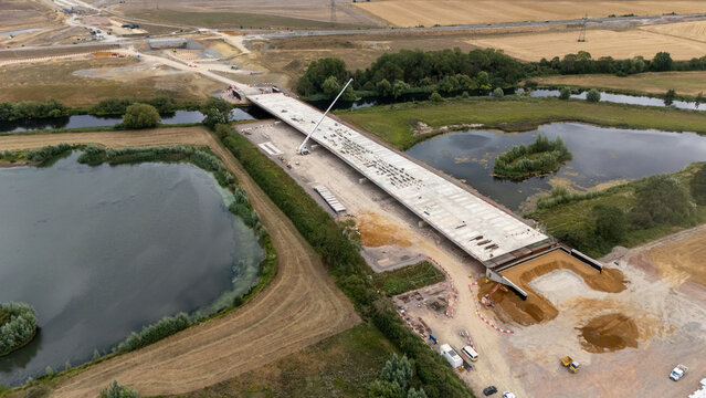 Aerial view of the Black Cat Roundabout bridge construction site featuring a concrete deck and a crane surrounded by rural fields and lakes in Chawston, England, United Kingdom.