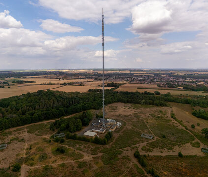 Aerial view of the Sandy Heath transmitting station, a tall mast rising above the surrounding rural landscape with fields, trees, and small buildings at its base in Sandy, England, United Kingdom.