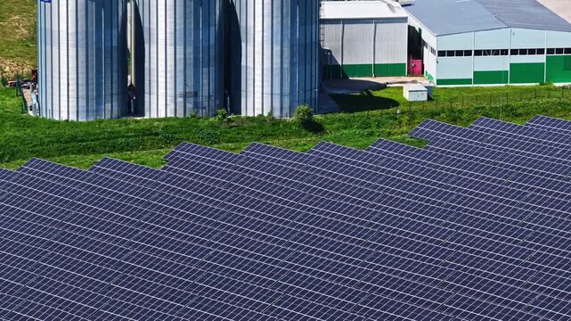 Aerial view of a large solar farm featuring rows of photovoltaic panels situated next to industrial grain silos and a warehouse facility in Riga, Latvia.