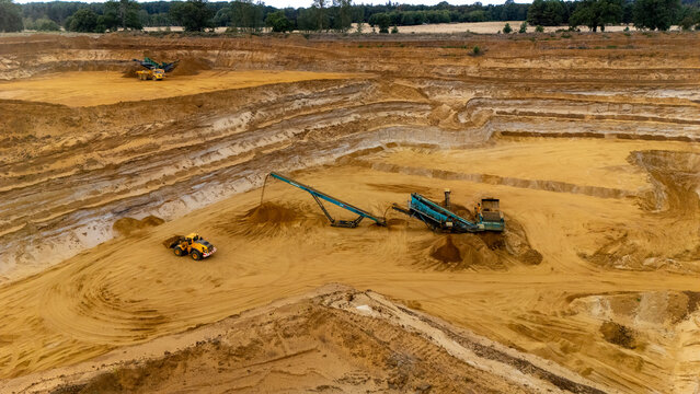 Aerial view of a sand quarry with heavy machinery, including an excavator and a conveyor belt, operating in a large open-pit mine in Sandy, England, United Kingdom.