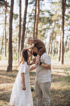 Happy family with young son on father's shoulders, parents kissing during summer walk in a green forest