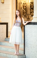 Fototapeta premium An elegant young woman in a stylish outfit poses on the stairs of a classic old building with columns. A fashion portrait, beauty, luxurious lifestyle, confidence, and urban elegance.