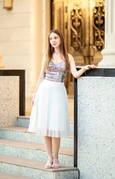 An elegant young woman in a stylish outfit poses on the stairs of a classic old building with columns. A fashion portrait, beauty, luxurious lifestyle, confidence, and urban elegance.