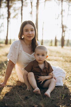 Mother and young son sit on grass in a sunlit pine forest, enjoying a summer evening in nature