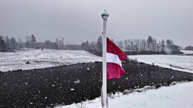 Aerial view of the national flag of Latvia waving on a flagpole over a snowy landscape with a frozen pond and trees in the background in Latvia.