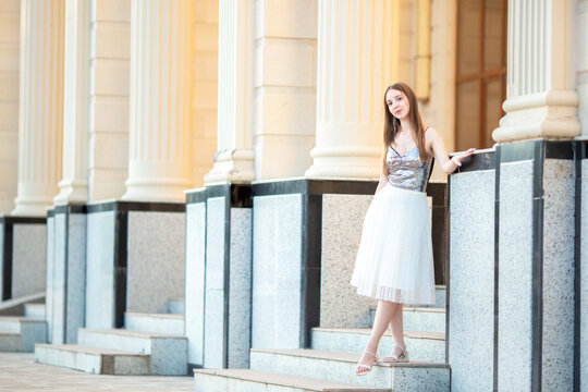 An elegant young woman in a stylish outfit poses on the stairs of a classic old building with columns. A fashion portrait, beauty, luxurious lifestyle, confidence, and urban elegance.