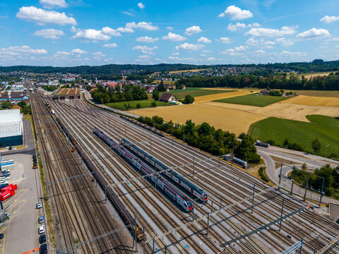 Aerial view of Daniken train station with passenger trains on multiple tracks, surrounded by green fields, rural landscape, and blue sky with clouds in Daniken, Solothurn, Switzerland.