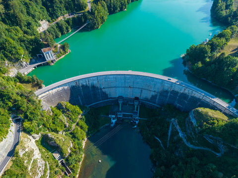 Aerial view of Barrage de Rossens, a concrete dam holding back turquoise reservoir waters, surrounded by dense forests and winding roads in Pont-la-Ville, Fribourg, Switzerland.