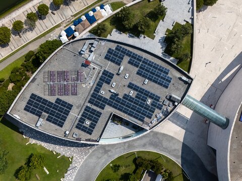 Aerial view of solar panels installed on a modern building roof surrounded by paved walkways and green landscaping in Lisboa, Lisboa, Portugal.