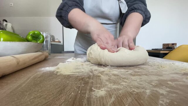 The hands of a female cook knead a lump of dough on a wooden table with white wheat flour.